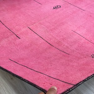 K&ouml;nnte beinhalten: Ein rosa Teppich mit schwarzen Linien und geometrischen Mustern. Der Teppich hat einen Fransenrand.