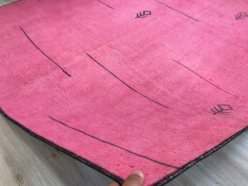 K&ouml;nnte beinhalten: Ein rosa Teppich mit schwarzen Linien und geometrischen Mustern. Der Teppich hat einen Fransenrand.