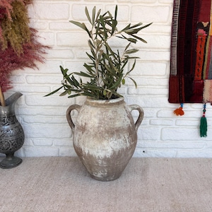 May include: A large, round, weathered ceramic vessel with two side handles and a distressed, mottled beige and white finish. The vessel contains a green olive branch with small leaves. To the left stands a smaller, ornate, dark metal pitcher.