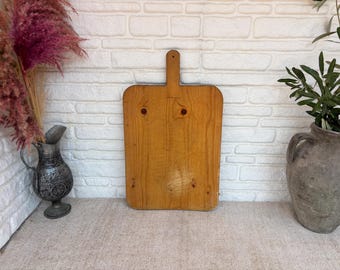 May include: A rectangular wooden cutting board with a vertical handle at the top center. The board features a natural light brown wood grain with visible knots and a smooth, flat surface, standing upright against a white textured brick wall.