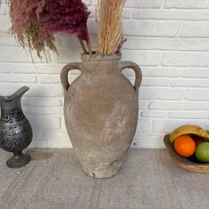 May include: A large, weathered, tan ceramic vase with two handles, filled with dried flowers. A decorative metal jug and a wooden bowl of fruit are also present.