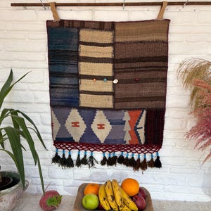 May include: A rectangular wall hanging tapestry featuring a patchwork of woven fabric panels in shades of blue, brown, tan, and orange. The lower section displays a geometric diamond pattern, finished with a fringe of dark tassels and small blue beads.