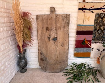 May include: A large, rectangular, weathered wooden board with a central handle at the top and a prominent knot hole in the middle. The wood displays a natural, light brown grain with visible signs of age, wear, and texture across its flat surface.