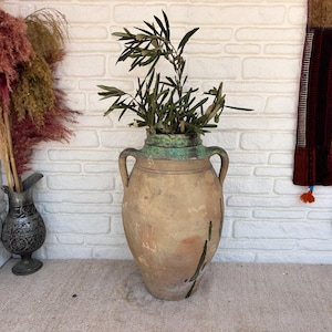 Puede incluir: Una gran ánfora de terracota desgastada con dos asas laterales y un distintivo borde esmaltado en verde azulado. El recipiente presenta una superficie texturizada de color marrón tierra con sutiles goteos verde oscuro y una rama de olivo.