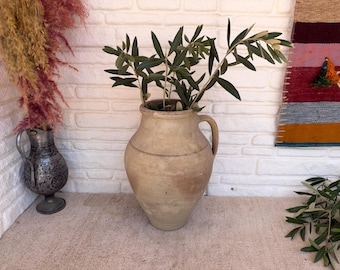 May include: A large, beige, weathered clay vessel with a single side handle, containing several green olive branches. The pot features a rounded body that tapers towards the base and a wide, open rim, showcasing a textured, matte, earthy finish.