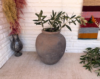 May include: A large, round, dark brown terracotta vessel with a wide opening and a thick rim. The vessel has a matte, weathered texture and contains several green olive branches with slender leaves extending from the top opening.