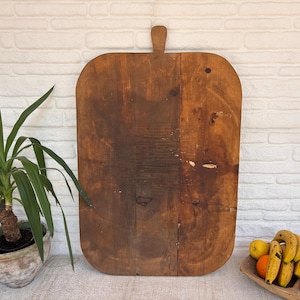 Peut inclure: Une grande planche à découper rectangulaire en bois avec une poignée. La planche est de couleur marron chaud avec des veines et des nœuds de bois visibles. Elle est présentée contre un mur de briques blanches, avec une plante en pot et un bol de fruits en bois au premier plan.