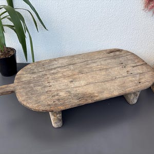 Puede incluir: Una tabla de servir rústica y desgastada de madera con un asa. La tabla rectangular está soportada por tres patas cortas y cilíndricas. La madera tiene un aspecto natural y envejecido con vetas y nudos visibles. La tabla se muestra sobre una superficie gris oscuro.