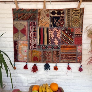 Può includere: Arazzo da parete rettangolare patchwork con una varietà di motivi geometrici nei toni del rosso, arancione, marrone e blu. Il tessuto è adornato con piccole nappe colorate lungo il bordo inferiore. È sospeso da un'asta di legno.