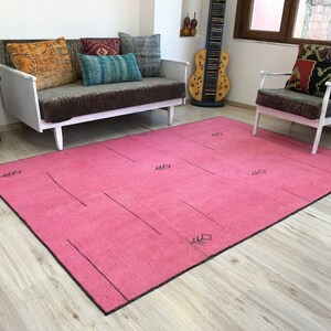 K&ouml;nnte beinhalten: Ein rosafarbener Teppich mit einem geometrischen Muster in Schwarz. Der Teppich liegt auf einem Holzboden und im Hintergrund befinden sich eine wei&szlig;e Couch und ein Stuhl.