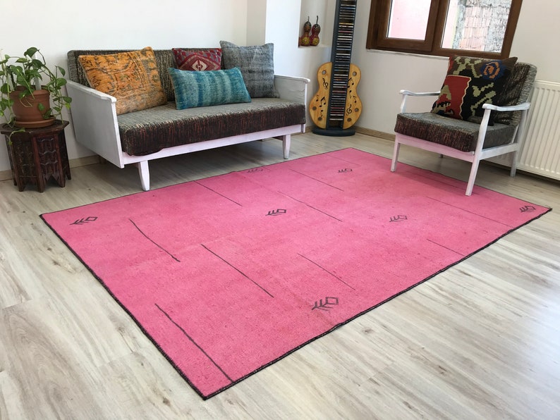 K&ouml;nnte beinhalten: Ein rosafarbener Teppich mit einem geometrischen Muster in Schwarz. Der Teppich liegt auf einem Holzboden und im Hintergrund befinden sich eine wei&szlig;e Couch und ein Stuhl.