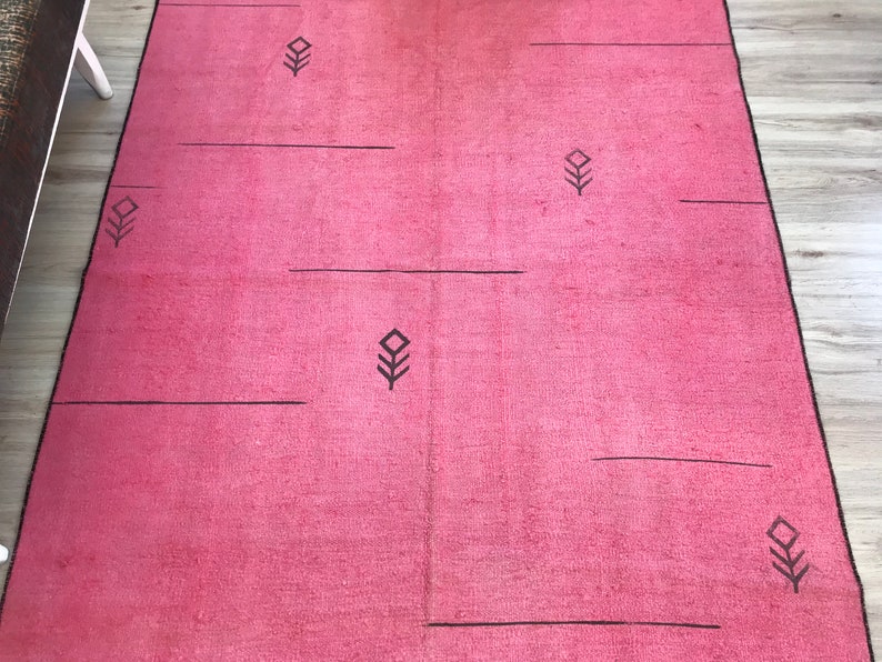 K&ouml;nnte beinhalten: Ein rosa Teppich mit einem geometrischen Muster aus schwarzen Linien und Diamanten. Der Teppich hat einen subtilen Ombre-Effekt, der von einem dunkleren Rosa oben zu einem helleren Rosa unten verblasst.
