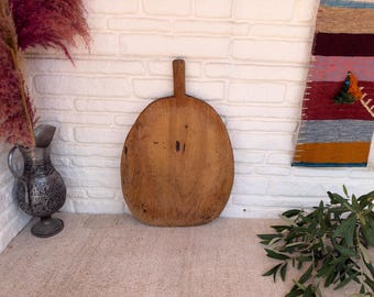 May include: A round, light brown wooden chopping board with a short, rectangular handle at the top. The surface of the board shows natural wood grain, visible wear, and small dark markings, indicating a textured and aged appearance.