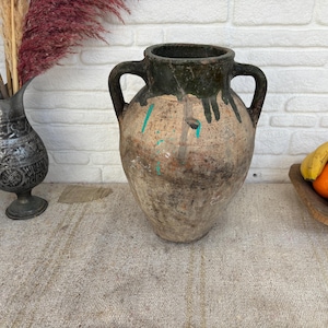 Pode incluir: Um grande recipiente de barro desgastado com duas alças, uma borda esmaltada em verde escuro com gotejamentos, um corpo texturizado em castanho claro e salpicos de tinta turquesa. O recipiente tem uma boca larga e base arredondada.