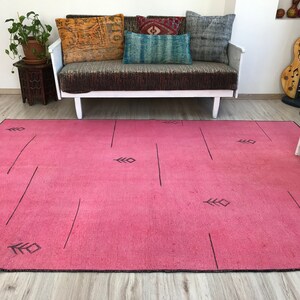 K&ouml;nnte beinhalten: Ein rosafarbener Teppich mit einem geometrischen Muster aus schwarzen Linien und Blattformen. Der Teppich liegt auf einem Holzboden vor einem wei&szlig;en Sofa mit Kissen.