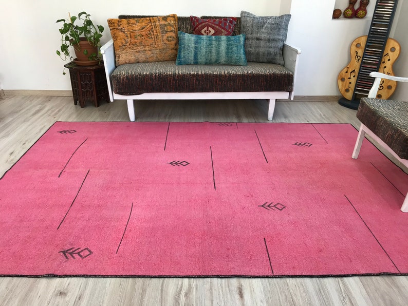 K&ouml;nnte beinhalten: Ein rosafarbener Teppich mit einem geometrischen Muster aus schwarzen Linien und Blattformen. Der Teppich liegt auf einem Holzboden vor einem wei&szlig;en Sofa mit Kissen.