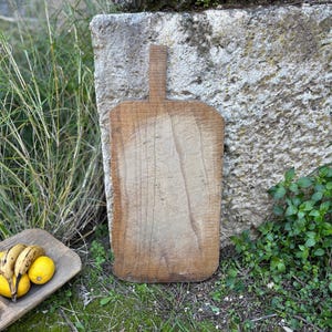 Puede incluir: Una tabla de cortar de madera rectangular grande con un asa. La tabla es de madera de color marrón claro y tiene vetas de madera visibles. Se apoya contra una pared de piedra, con un cuenco de madera con fruta en primer plano.