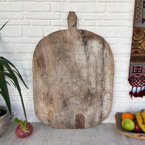 Puede incluir: Una tabla de cortar de madera grande y desgastada con forma redondeada y un pequeño mango en la parte superior. La madera presenta una veta natural de color marrón claro con signos visibles de uso, incluyendo arañazos y una mancha oscura.