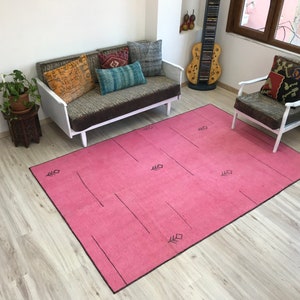 K&ouml;nnte beinhalten: Ein rosafarbener Teppich mit einem geometrischen Muster in Schwarz. Der Teppich liegt auf einem hellen Holzboden und im Hintergrund befinden sich ein wei&szlig;es Sofa und ein wei&szlig;er Sessel.