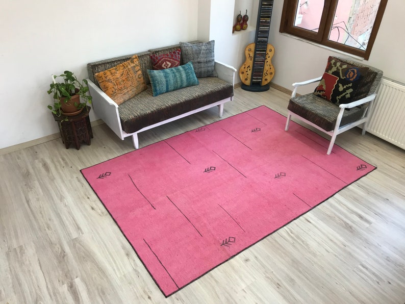 K&ouml;nnte beinhalten: Ein rosafarbener Teppich mit einem geometrischen Muster in Schwarz. Der Teppich liegt auf einem hellen Holzboden und im Hintergrund befinden sich ein wei&szlig;es Sofa und ein wei&szlig;er Sessel.