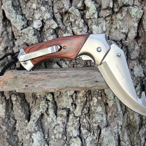 May include: A folding pocket knife with a wooden handle and a stainless steel blade. The blade has a curved edge and a hook at the end. The handle has a metal clip. The knife is resting on a small wooden branch.
