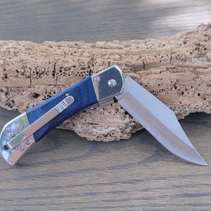 May include: A blue and silver pocket knife with a wooden handle. The blade is open and the knife is resting on a piece of driftwood.