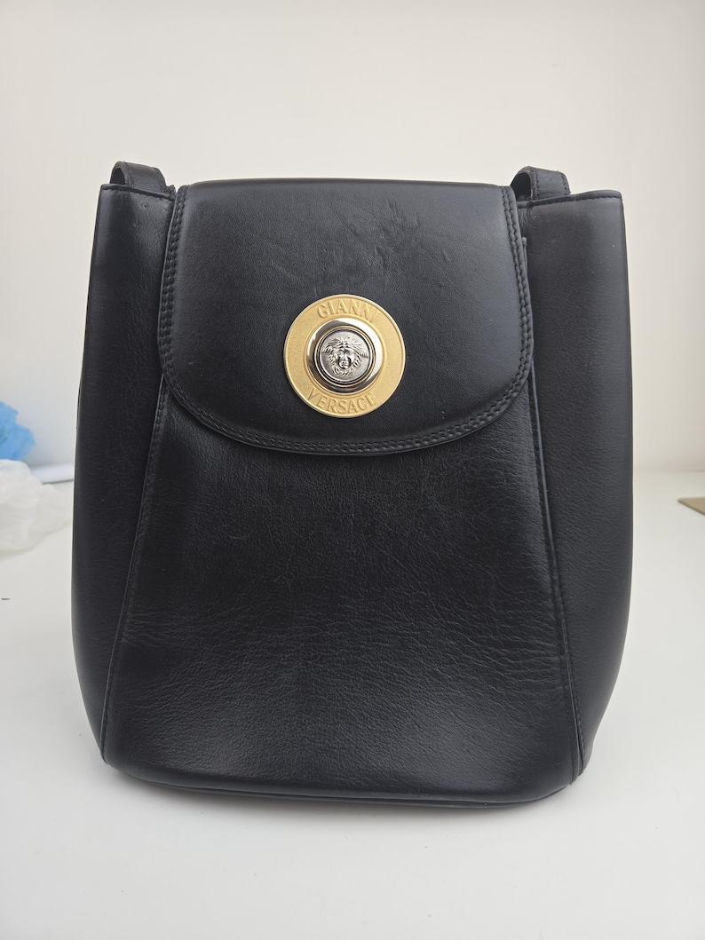Pu&ograve; includere: Borsa a tracolla in pelle nera con chiusura a patta. La borsa presenta un emblema circolare dorato con la scritta "GIANNI VERSACE" e una testa di Medusa. La borsa ha una forma strutturata e un manico superiore.