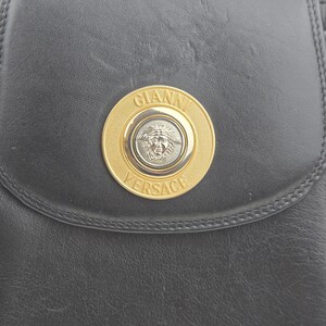 Pu&ograve; includere: Borsa a tracolla in pelle nera con un emblema Gianni Versace dorato e argentato. L'emblema circolare presenta il nome del marchio e una testa di Medusa. La borsa ha una chiusura a patta e cuciture.