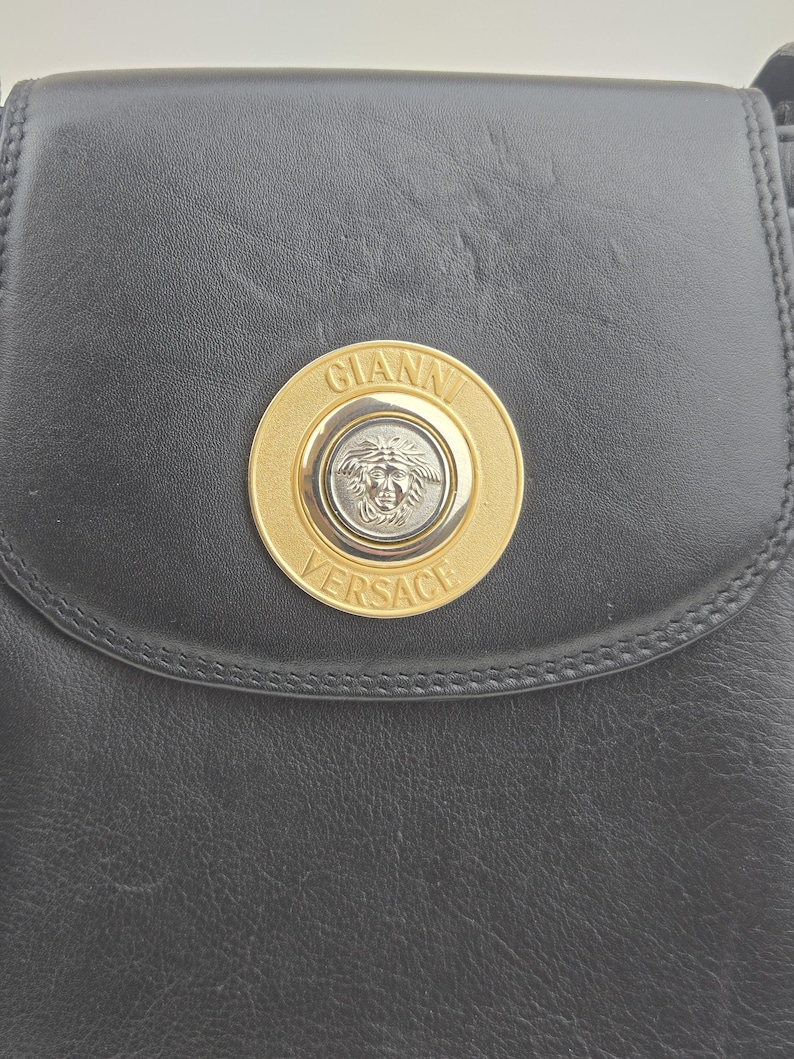 Pu&ograve; includere: Borsa a tracolla in pelle nera con un emblema Gianni Versace dorato e argentato. L'emblema circolare presenta il nome del marchio e una testa di Medusa. La borsa ha una chiusura a patta e cuciture.