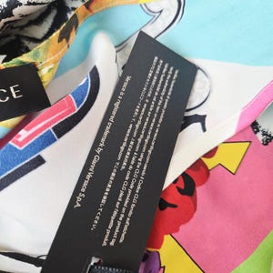 Puede incluir: Una prenda de tela multicolor con un estampado abstracto vibrante en tonos rosa, amarillo, azul y negro. Se adjunta una etiqueta rectangular negra que muestra texto blanco con instrucciones para verificar la autenticidad en versace.certilogo.com.