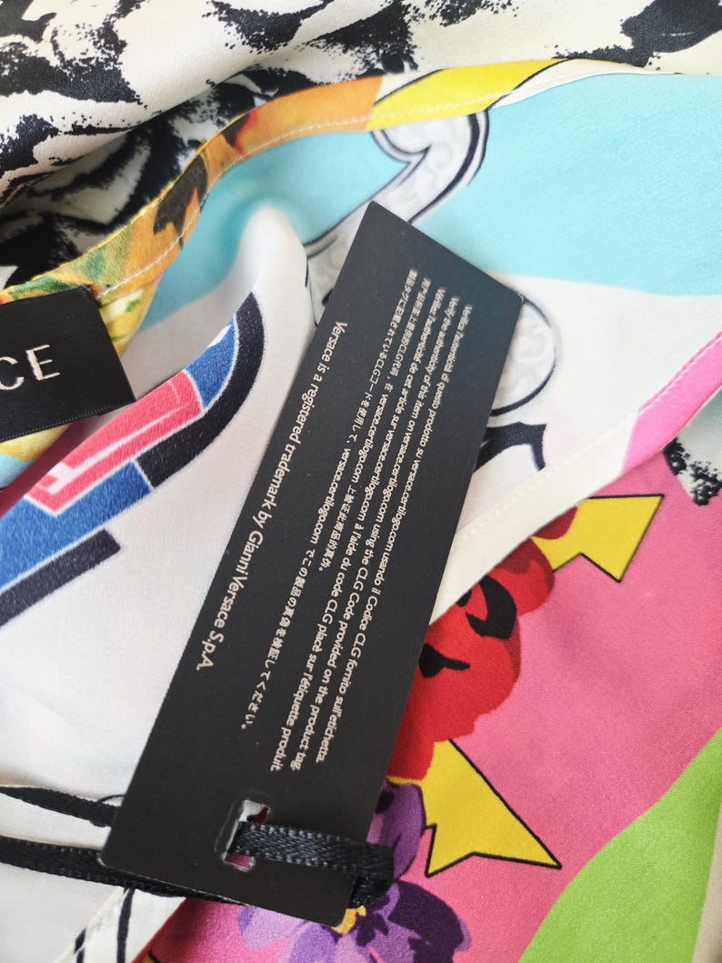 Puede incluir: Una prenda de tela multicolor con un estampado abstracto vibrante en tonos rosa, amarillo, azul y negro. Se adjunta una etiqueta rectangular negra que muestra texto blanco con instrucciones para verificar la autenticidad en versace.certilogo.com.