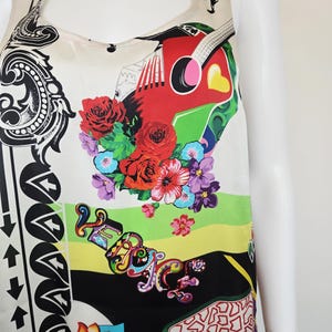 Puede incluir: Una blusa sin mangas con un estampado gr&aacute;fico vibrante y multicolor sobre un fondo crema. El dise&ntilde;o incluye rosas rojas, una guitarra estilizada, motivos florales y la palabra VERSACE en letras decorativas coloridas, junto a patrones de flechas negras.