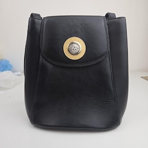 Pu&ograve; includere: Borsa a tracolla in pelle nera con chiusura a patta. La borsa presenta un emblema circolare dorato con le parole "GIANNI VERSACE" in un cerchio dorato. La borsa ha una forma rettangolare e un manico superiore.