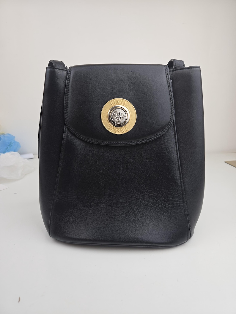 Pu&ograve; includere: Borsa a tracolla in pelle nera con chiusura a patta. La borsa presenta un emblema circolare dorato con le parole "GIANNI VERSACE" in un cerchio dorato. La borsa ha una forma rettangolare e un manico superiore.