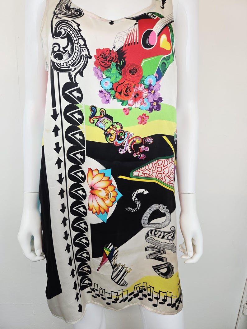 Puede incluir: Un vestido sin mangas con base color crema, negro y amarillo con un estampado gr&aacute;fico vibrante. El dise&ntilde;o incluye una guitarra roja, rosas coloridas, un zapato de tac&oacute;n, notas musicales, una flor de loto y el texto "VERSACE" en letras estilizadas.