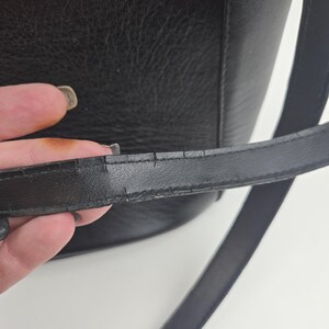Pu&ograve; includere: Primo piano di una borsa in pelle nera. La borsa ha una superficie strutturata e una tracolla lunga e sottile. La tracolla mostra segni di usura. Il design della borsa &egrave; semplice e classico, adatta all'uso quotidiano.