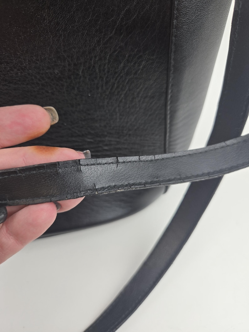 Pu&ograve; includere: Primo piano di una borsa in pelle nera. La borsa ha una superficie strutturata e una tracolla lunga e sottile. La tracolla mostra segni di usura. Il design della borsa &egrave; semplice e classico, adatta all'uso quotidiano.