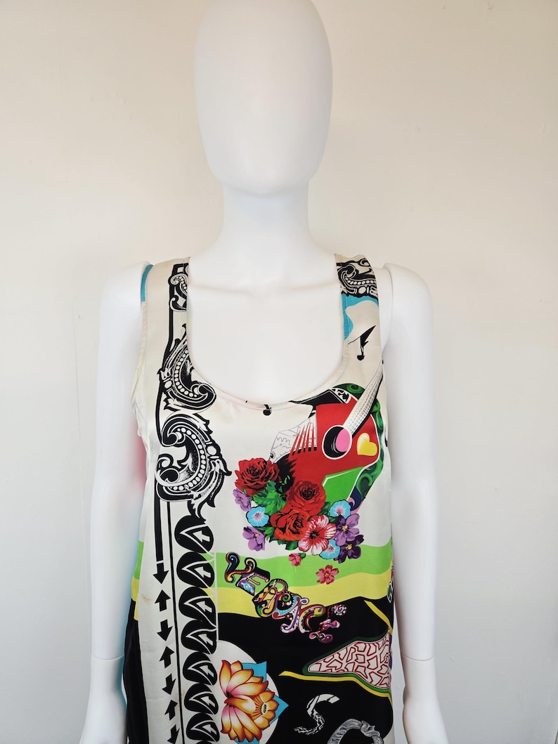 Puede incluir: Una camiseta sin mangas con base color crema y un estampado gr&aacute;fico vibrante. El dise&ntilde;o incluye una guitarra roja, arreglos florales coloridos, bordes con volutas negras y la palabra Versace en letras estilizadas sobre una franja verde brillante.