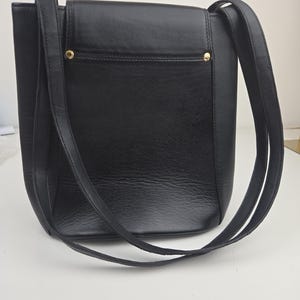 Pu&ograve; includere: Una borsa a tracolla in pelle nera con forma rettangolare e lunghe cinghie. La borsa presenta una chiusura a patta e dettagli metallici color oro. La borsa &egrave; progettata con un'estetica minimalista.