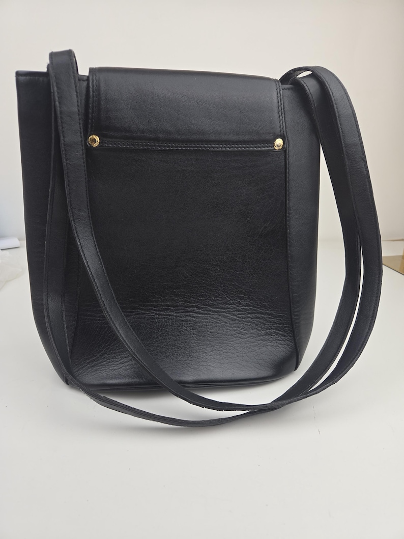 Pu&ograve; includere: Una borsa a tracolla in pelle nera con forma rettangolare e lunghe cinghie. La borsa presenta una chiusura a patta e dettagli metallici color oro. La borsa &egrave; progettata con un'estetica minimalista.
