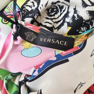 Puede incluir: Una prenda de Versace con una etiqueta de tela negra que muestra el nombre de la marca y el logotipo de Medusa. El material presenta un estampado gr&aacute;fico vibrante y multicolor que incluye patrones florales en blanco y negro, adem&aacute;s de formas abstractas en rosa, verde y azul.