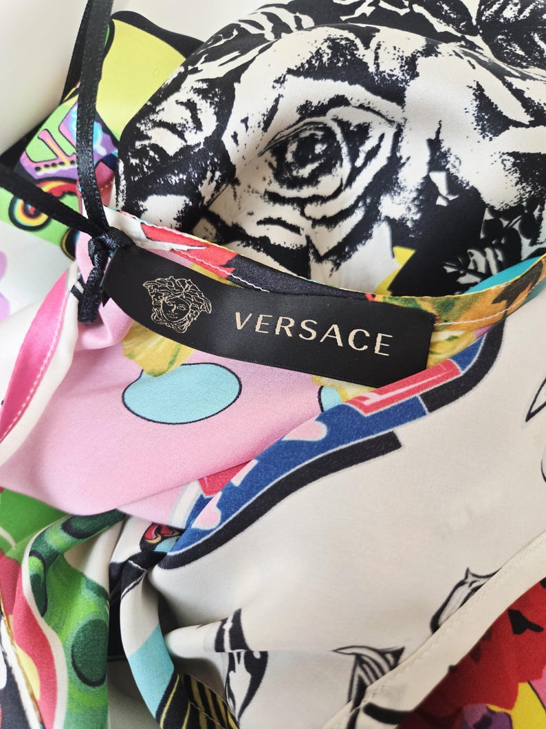 Puede incluir: Una prenda de Versace con una etiqueta de tela negra que muestra el nombre de la marca y el logotipo de Medusa. El material presenta un estampado gr&aacute;fico vibrante y multicolor que incluye patrones florales en blanco y negro, adem&aacute;s de formas abstractas en rosa, verde y azul.