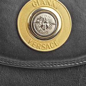 Pu&ograve; includere: Primo piano di una borsa in pelle nera con un emblema Gianni Versace oro e argento. L'emblema &egrave; circolare, con il nome del marchio e un disegno a testa di Medusa.