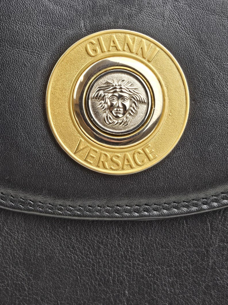Pu&ograve; includere: Primo piano di una borsa in pelle nera con un emblema Gianni Versace oro e argento. L'emblema &egrave; circolare, con il nome del marchio e un disegno a testa di Medusa.