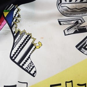 Puede incluir: Un pa&ntilde;uelo de seda con fondo blanco y patrones gr&aacute;ficos en negro, amarillo y varios colores. El dise&ntilde;o incluye formas geom&eacute;tricas estilizadas en blanco y negro, una secci&oacute;n triangular amarilla y letras negras con un estampado tipo cebra.