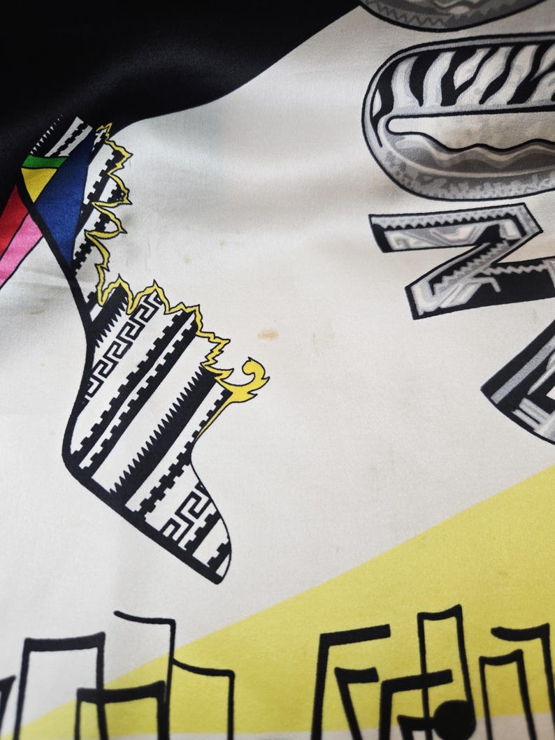 Puede incluir: Un pa&ntilde;uelo de seda con fondo blanco y patrones gr&aacute;ficos en negro, amarillo y varios colores. El dise&ntilde;o incluye formas geom&eacute;tricas estilizadas en blanco y negro, una secci&oacute;n triangular amarilla y letras negras con un estampado tipo cebra.