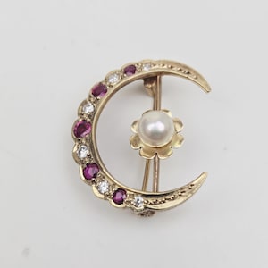 Puede incluir: Broche dorada con forma de media luna, con una perla central y un adorno floral. La luna está adornada con piedras preciosas rojas y claras alternas. Una joya clásica.