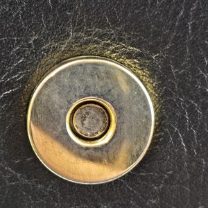 Pu&ograve; includere: Primo piano di una chiusura magnetica su pelle nera. La chiusura &egrave; circolare con un anello esterno argentato e un anello interno dorato. La pelle ha un aspetto strutturato.
