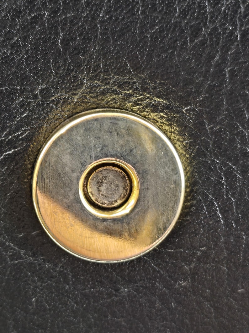 Pu&ograve; includere: Primo piano di una chiusura magnetica su pelle nera. La chiusura &egrave; circolare con un anello esterno argentato e un anello interno dorato. La pelle ha un aspetto strutturato.