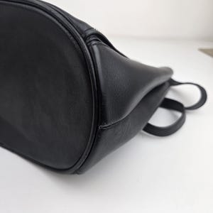 Pu&ograve; includere: Primo piano di una borsa in pelle nera. La borsa ha un fondo arrotondato e una consistenza liscia. Sono visibili due piccole maniglie. La borsa sembra vuota ed &egrave; posta su uno sfondo bianco.